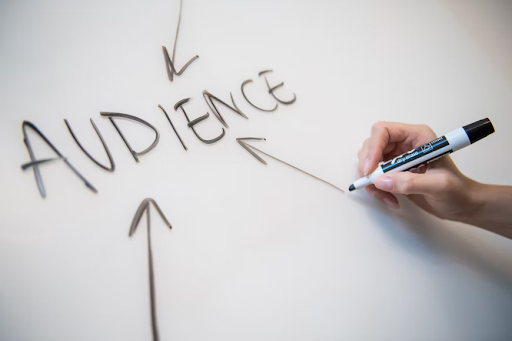 Close-up photograph of a whiteboard with the word 'audience' written in bold letters at the center. Several arrows, drawn in different colors, point towards the word, indicating its importance. The arrows symbolize the direction of attention and highlight the central focus of understanding and engaging with the target audience.