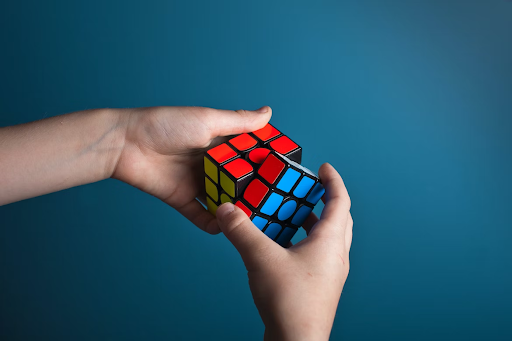 A pair of hands solving a Rubik's Cube. The hands are positioned around the Rubik's Cube, twisting and turning its colorful sides to solve the puzzle. The hands appear focused and skillful, demonstrating dexterity and precision as they manipulate the cube. The Rubik's Cube itself consists of multiple small, colored squares arranged in a 3x3 grid, forming a larger cube shape. The colors on the cube's faces include red, blue, green, orange, yellow, and white, each representing a different side of the puzzle.