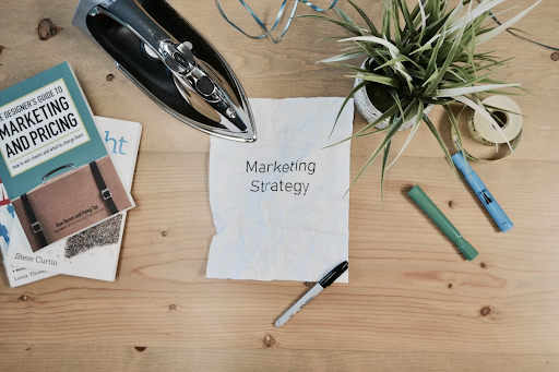 Image of an office desk neatly arranged with various items related to marketing. A laptop is positioned in the center, indicating work in progress. Surrounding the laptop are marketing books, pens, and other stationery, suggesting a professional environment. Additionally, there is a paper prominently placed on the desk, displaying the words 'Marketing Strategy' written on it, indicating the ongoing planning and development of a strategic approach.