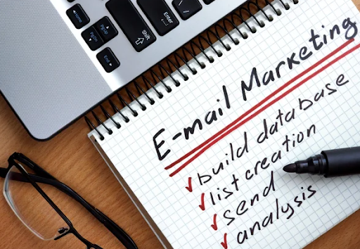 : A laptop and a notebook displaying "Email Marketing: Build Database, List Creation, Send, Analysis". The alt text describes a laptop and a notebook placed side by side. The laptop screen is not specified in detail, while the notebook has the text "Email Marketing: Build Database, List Creation, Send, Analysis" written on it. The notebook highlights key components of email marketing, including building a database, creating lists, sending emails, and conducting analysis. The laptop and notebook combination suggests a workspace or environment dedicated to email marketing tasks and activities.