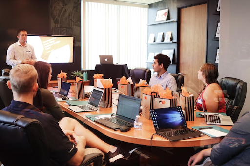A conference room with multiple people seated around a long table, engaged in a lively discussion. Each person has a laptop in front of them, with various documents and presentations displayed on the screens. The room is well-lit and spacious, with large windows providing a view of the city skyline in the background. The participants appear attentive, gesturing and exchanging ideas, creating a collaborative and productive atmosphere.